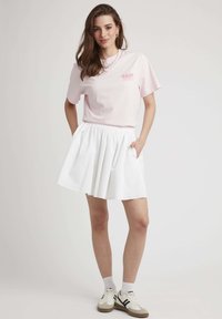 T-shirt en coton rose avec "RASP" imprimé en blanc sur la poitrine, associé à une jupe plissée blanche et des baskets légères avec des accents noirs.