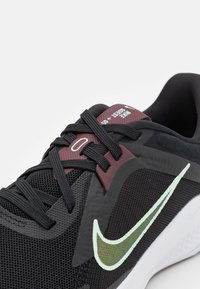 Black athletic shoe with a textured mesh upper, maroon accents, black laces, and a reflective mint green logo on the side.