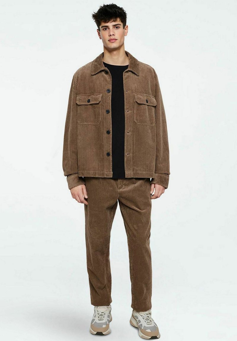 Veste en velours côtelé marron et pantalon assorti, avec deux poches poitrine, fermeture à bouton et tissu texturé. Porté par-dessus une chemise noire.