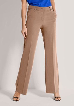 Frau trägt beige, hochgeschnittene, weit geschnittene Hose mit Taschen, kombiniert mit schwarzen Sandalen mit offenen Zehen und einem blauen Oberteil.