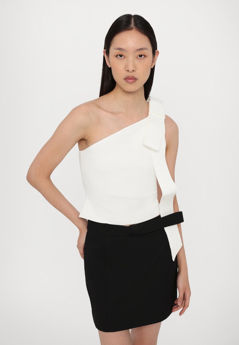 Asymmetrical white top with a wide strap and bow accent, paired with a black fitted skirt featuring a matching belt.
