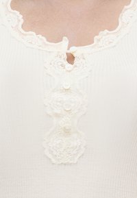 Κοντινό πλάνο από κορυφή ριμπ ύφανσης σε κρεμ χρώμα με δαντελένιο τελείωμα στο λαιμό και διακοσμητική κάθετη φαρδιά δαντέλα με μικρά κουμπιά.