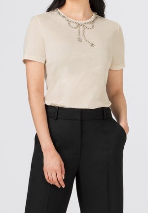 T-shirt beige clair à manches courtes avec un col arrondi, orné d'un détail en chaîne argentée décorative. Assorti à un pantalon noir ajusté.