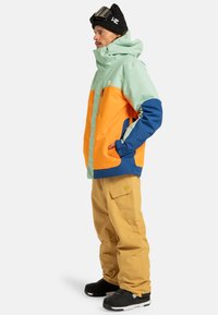 Giacca da snowboard colorata con pannelli verde chiaro, arancione e blu; design con cappuccio e tasche con zip. Abbinata a pantaloni cargo beige e scarpe nere.