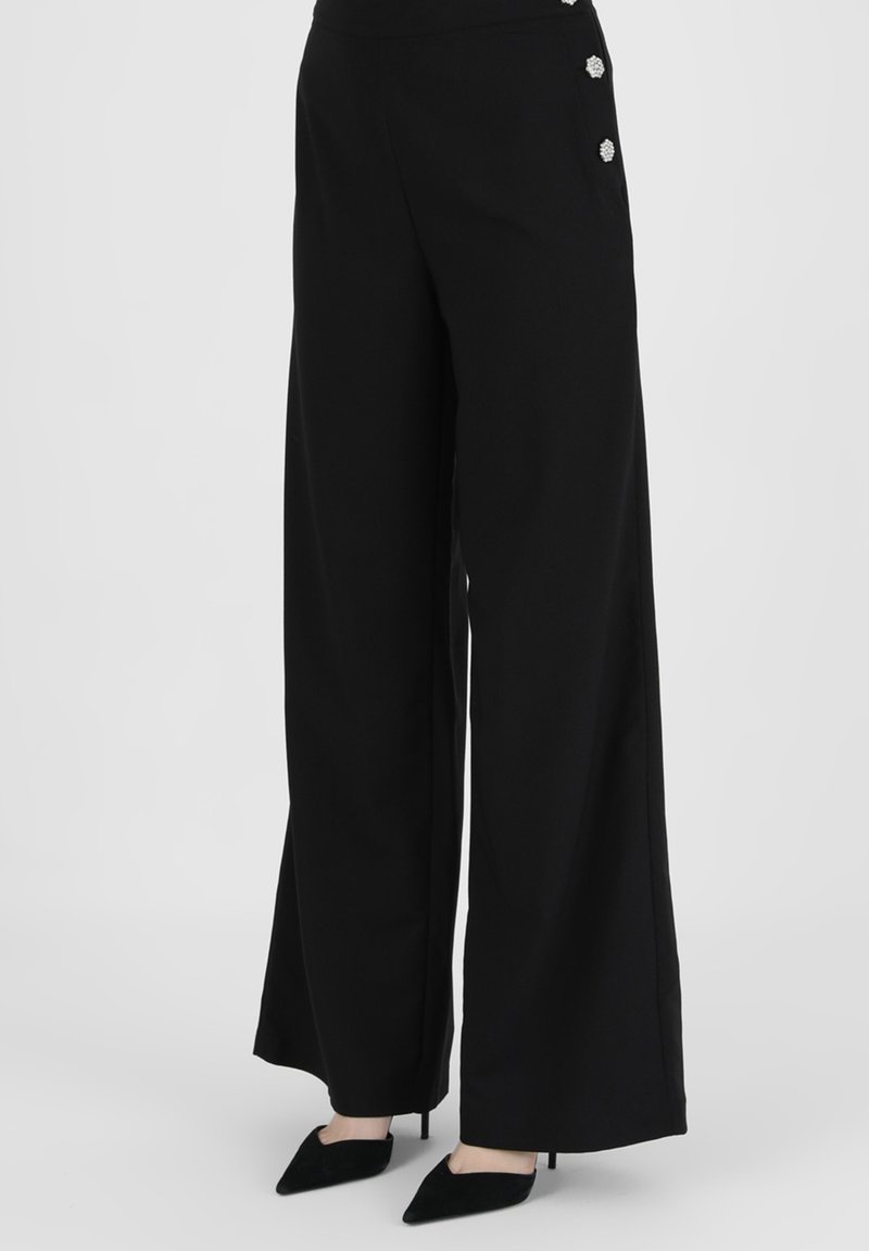 Pantalon noir à jambes larges, taille haute avec boutons décoratifs, porté avec des escarpins noirs à bout pointu sur fond uni.