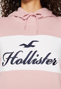 Zblízka růžová a bílá mikina s kapucí se staženou šňůrkou a černým vyšitým logem Hollister a ptáčkem.