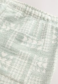 Tessuto strutturato in verde chiaro con motivi floreali e geometrici bianchi, dotato di una vita elastica arricciata. Materiale morbido e intrecciato.