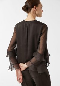 Blouse noire transparente avec manches longues superposées et à volants, encolure ronde et dos lisse. Le tissu semble léger et légèrement texturé.