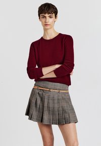 Un pull en maille bordeaux avec un col rond associé à une jupe plissée à carreaux gris avec une ceinture beige. Texture lisse et design structuré.