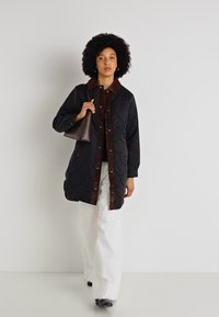 Kvadrični črni plašč z rjavim kordnim ovratnikom in sprednjim plaščem; model drži rjav nahrbtnik; v kombinaciji z belimi širokimi hlačami.