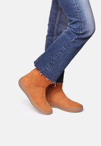 Pies que llevan botas de ante color marrón claro hasta el tobillo con cremalleras laterales, combinadas con jeans de mezclilla azul con dobladillos deshilachados sobre un fondo blanco.