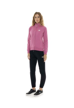 Une femme porte une veste rose zippée, un pantalon de jogging noir et des baskets noires sur un fond blanc.