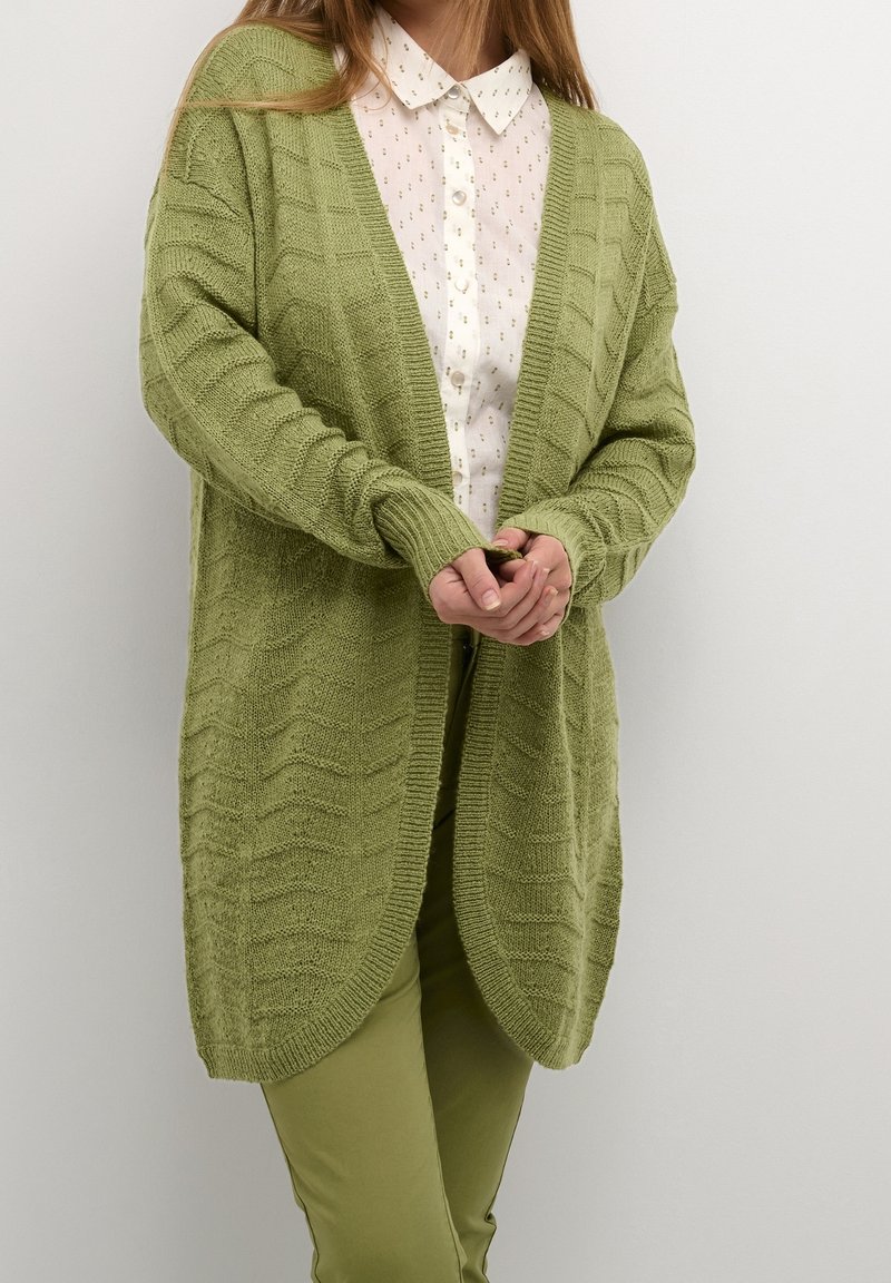 Femme portant un cardigan en tricot vert clair sur une chemise blanche boutonnée et un pantalon vert clair, debout devant un fond uni.