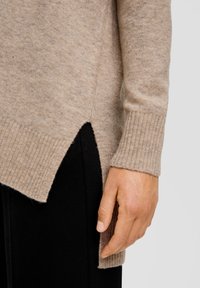 Beige gebreide trui met geribbelde manchetten en zijsplits, afgebeeld met een hand die de zoom aanpast. Gladde textuur, casual stijl en een getailleerde pasvorm.