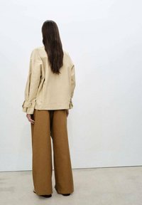 Jachetă oversized bej cu mâneci largi, purtată împreună cu pantaloni largi maronii. Textură fină și design minimalist pe un fundal neutru.