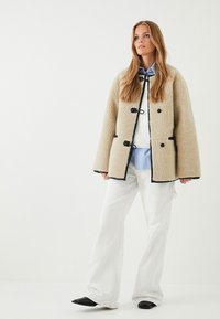 Chaqueta de forro polar beige con ribete negro y cierres de botonadura, llevada sobre un suéter blanco y una camisa azul claro, combinada con pantalones anchos blancos.