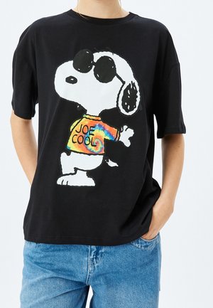 Osoba nosząca czarną koszulkę z dużą kreskówką Snoopy w okularach przeciwsłonecznych i kolorową koszulką "Joe Cool", połączoną z niebieskimi dżinsami.