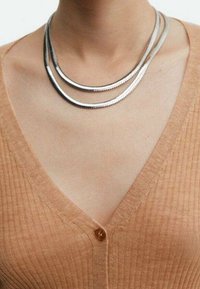 Stříbrný vícevrstvý náhrdelník s hladkými, texturovanými prameny, který je nošený přes žebrovaný, svetrový kardigán světle hnědé barvy s výstřihem do V a knoflíkovým detalem.