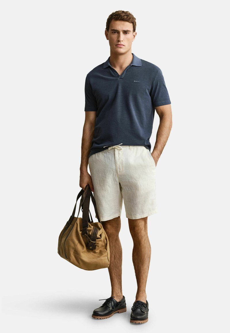 Mann, der ein marineblaues Poloshirt und beige Shorts trägt, eine braune Reisetasche hält, mit einer Hand in der Tasche steht und einen neutralen Gesichtsausdruck zeigt.