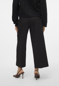 Pantalon large noir avec une texture lisse et des plis à l'avant, associé à des mules à talons bruns. Vue de derrière, modèle portant un pull noir.