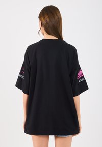 Tricou oversized negru din bumbac, cu mâneci scurte. Mânecile din stânga au un text roz și un design grafic, cu o textură netedă.