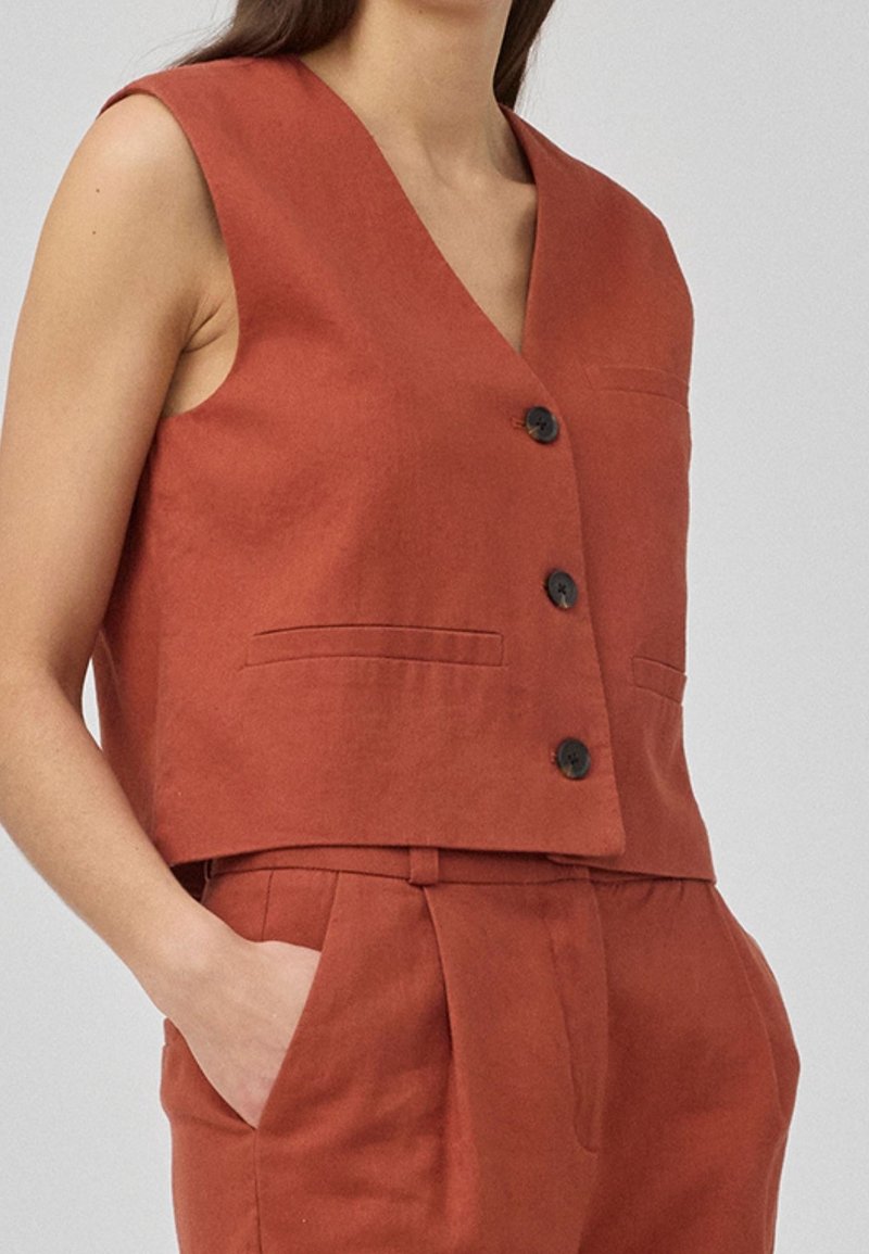 Gilet sans manches couleur rouille à boutons avec deux poches avant, porté avec un pantalon taille haute assorti, les mains dans les poches.