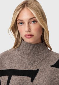 Maglione a maglia marrone con collo alto, caratterizzato da un motivo grafico nero. Tessuto morbido con vestibilità comoda e dettagli di cucitura visibili.