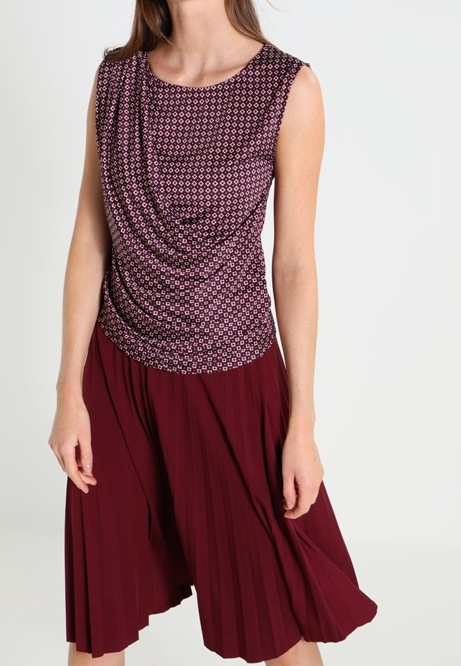 Femme portant un haut sans manches à motifs et une jupe plissée bordeaux, debout devant un fond clair uni.