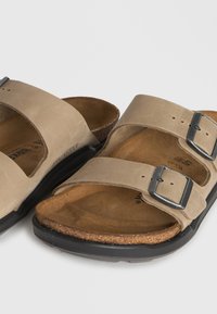 Μπεζ σουέντ σανδάλια Birkenstock με δύο ρυθμιζόμενες μεταλλικές αγκράφες και καμπυλωτές φελντιμπέτες φτιαγμένες από φελλό σε μαύρες λαστιχένιες σόλες.