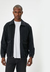 Hombre vestido con una chaqueta de mezclilla negra sobre una camiseta blanca y jeans negros, de pie contra un fondo blanco liso.