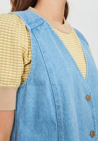 Denimweste in Hellblau mit Knopfschluss, breiten Schulterträgern und V-Ausschnitt. Über einem strukturierten gelben Oberteil getragen.