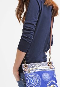 Blaue Schultertasche mit geometrischen Mustern, Leder-Schulterriemen und metallischen Beschlägen. Material aus Canvas mit strukturierten Akzenten.