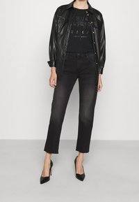 Camisa chaqueta de piel sintética negra con botones a presión, superpuesta sobre una camiseta negra con texto de strass, combinada con jeans negros recortados y tacones.