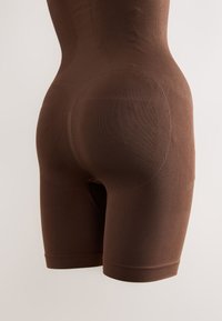 Shorts moulants marron sans couture sur mannequin, s'étendant jusqu'au milieu de la cuisse avec des zones de contour pour l'amélioration des fessiers.