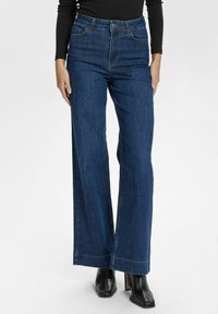 Wide-leg blue denim jeans with high waist, visible stitching, and a polished finish. Paired with black heeled ankle boots.