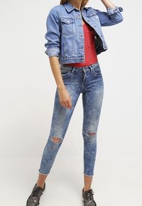 Světle modrá riflová bunda s zkráceným střihem a dvěma kapsami, doplněná červeným tílkem a roztrhanými světle modrými džíny, obutá s černými teniskami.