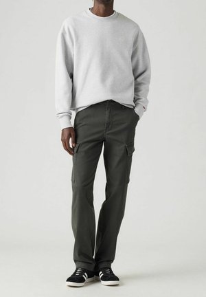 Homme portant un sweat-shirt gris clair à manches longues et un pantalon cargo vert foncé avec des baskets noires, debout devant un fond blanc.