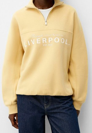Person wearing a beige quarter-zip sweatshirt with "Merseyside, United Kingdom LIVERPOOL EST. 2024" text and dark blue jeans.