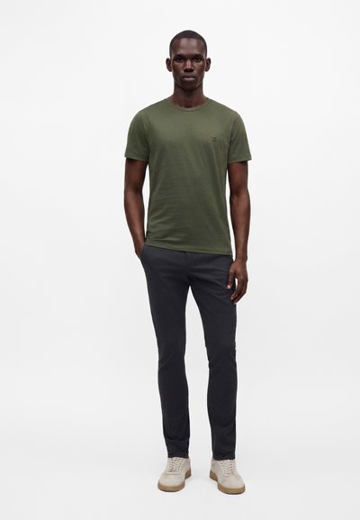 Homme debout, les mains dans les poches, portant un t-shirt vert olive, un pantalon foncé et des baskets beiges sur fond blanc.