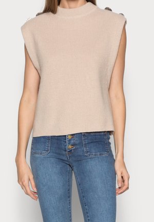 Pull tricot beige sans manches avec des détails de boutons aux épaules, porté avec un jean bleu taille haute avec boutons apparents et poches avant.