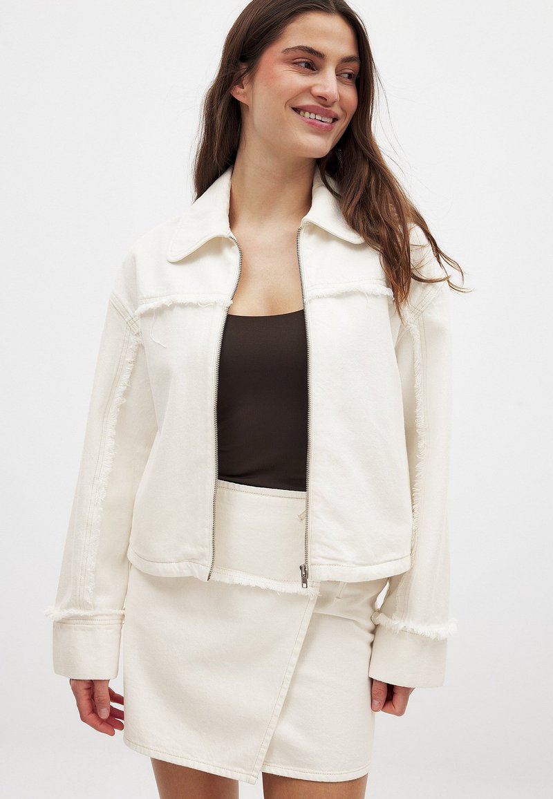 Valge denimpusa, millel on tõmblukuga esiosa, teravad kraed ja narmastega ääred, kombineeritud sobiva sätendava seeliku ja lendava tekstuuriga. Sellel on sile pind ja juhuslik lõige.