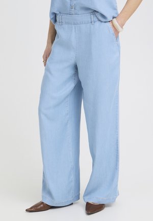 Kobieta w jasnoniebieskich szerokich spodniach z rękami w kieszeniach, w połączeniu z brązowymi spiczastymi butami i jasnoniebieską bluzką.