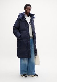 Giacca puffer blu marina con cappuccio, caratterizzata da un design trapuntato, due tasche laterali e una zip frontale. Abbinata a una felpa grigia chiara e jeans larghi.