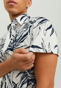 Camicia a maniche corte in cotone bianco con motivo di foglie blu, design con bottoni e colletto strutturato. Presenta polsini arrotolati.