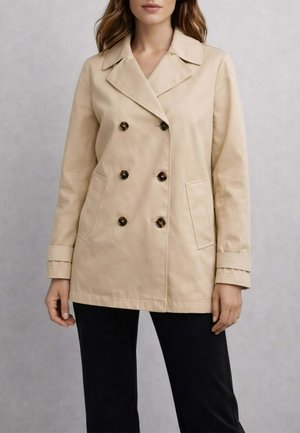 Donna con giacca doppiopetto beige con colletto a rever e bottoni in tartaruga, abbinata a pantaloni neri, in piedi davanti a uno sfondo neutro.