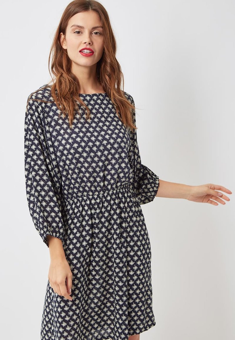 Mujer con cabello largo castaño que lleva un vestido azul marino con patrones geométricos blancos y mangas tres cuartos, de pie frente a un fondo claro y liso.