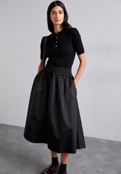 Top corto a maniche corte in riga nera con bottoni, abbinato a una gonna maxi in raso nera con vita elastica e tasche. Scarpe nere completano il look.