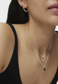 Collares de plata en capas con un colgante negro, que muestran una textura suave. Pequeños pendientes de aro complementan el diseño simple y moderno.