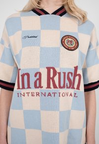 Person iført en kortærmet skjorte i blå og beige tern med teksten "In a Rush International" og et rundt mærke på brystet.