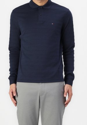 Homme portant un polo bleu marine à manches longues avec un petit logo rouge, blanc et bleu sur la poitrine, associé à un pantalon gris clair.